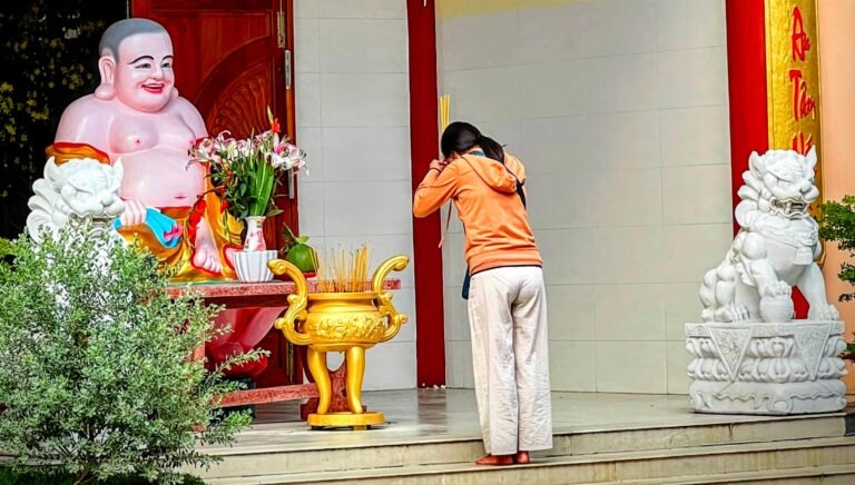 Woman bowing in prayer beside a laughing Buddha statue and a stone lion at a Vietnamese temple