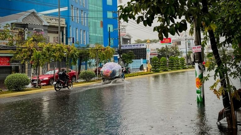 Flooded street in Soc Trang during rainy season, with a motorbike and car wading through standing water